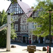 Skulptur Der Hungerbrunnen Skulptur eines überdimensionalen Nagels vor Fachwerkhäusern im Sonnenschein, umgeben von Bäumen.