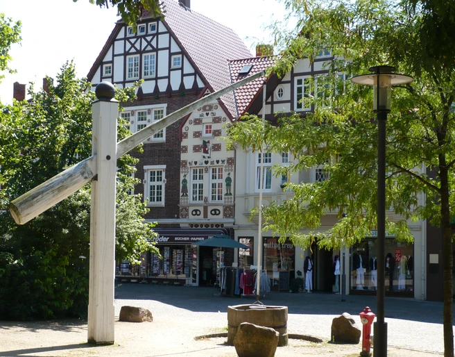 Skulptur Der Hungerbrunnen Skulptur eines überdimensionalen Nagels vor Fachwerkhäusern im Sonnenschein, umgeben von Bäumen.