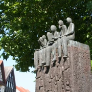 Skulptur "Der Vorleser" Skulptur von sitzenden Figuren auf einem hohen Sockel, umgeben von grünen Blättern und Fachwerkhäusern.