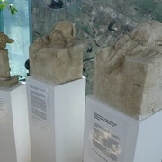 Skulptur Epitaph Drei abstrakte Skulpturen auf weißen Sockeln vor einer Karte zeigen unterschiedliche menschliche Kopfformen.