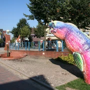 Bunte Skulptur eines Aals auf einem Gehweg vor traditioneller Backsteinarchitektur und blauem Himmel.