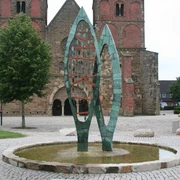 Brunnenskulptur STATIO Skulptur aus grünem Metall, bestehend aus zwei geschwungenen Formen, auf einem gepflasterten Platz.