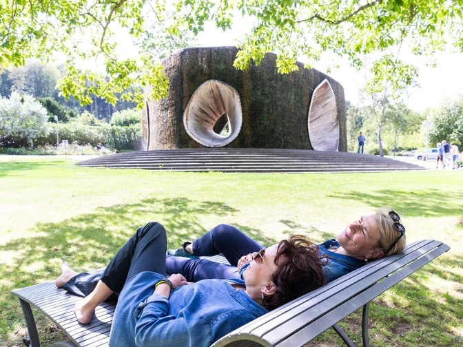 SoleArena Bad Essen Zwei Frauen entspannen auf einer Bank vor einem großen, skulpturalen Brunnen im Park.