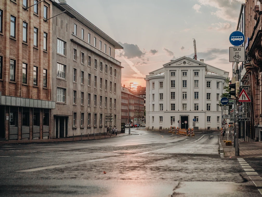 Blick über eine breite, nasse und von mehrstöckigen Gebäuden begleitete Straße auf das Theater Aachen und den Sonnenuntergang im Hintergrund.