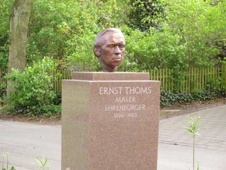 Ernst Thoms-Bronzekopf Eine Bronzebüste von Ernst Thoms steht auf einem steinernen Sockel in einer grünen Parkanlage.