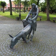 Skulptur Esel und Mönch Skulptur eines Mönchs auf einem bückenden Esel inmitten eines gepflasterten, von Bäumen umgebenen Platzes.