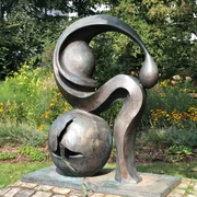 Eine Bronze-Skulptur mit abstrakten Formen steht vor einer blühenden Gartenkulisse.