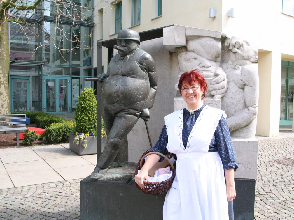 Kinderführung mit Anna - Frau vom Zigarrenmacher Frau in traditioneller Kleidung steht lächelnd neben einer Bronzeskulptur in einem belebten Innenhof.