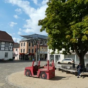 Achimer Glockenspiel Auf einem sonnigen Marktplatz in Achim sitzt eine Person neben einem roten Holzspielzeugauto unter einem Baum.