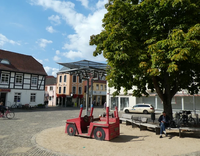 Achimer Glockenspiel Auf einem sonnigen Marktplatz in Achim sitzt eine Person neben einem roten Holzspielzeugauto unter einem Baum.
