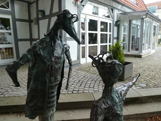 Skulptur "Komm" Zwei metallische Skulpturen mit menschlichen Zügen stehen vor einem Fachwerkgebäude mit Kieselsteinen.