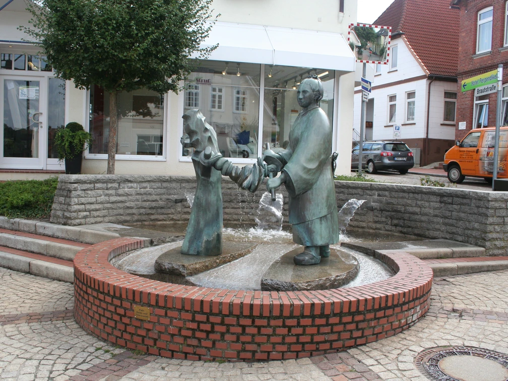 Brunnen - Die Wringerin Brunnen-Skulptur einer Frau, die Wäsche auswringt, inmitten eines gepflasterten Platzes mit Bäumen.