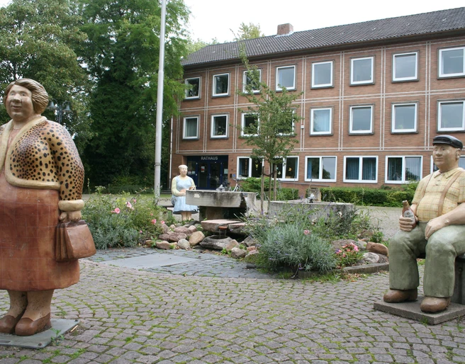Rathaus Petershagen mit Lechner Figuren Bronzefiguren von Menschen vor dem Rathaus Petershagen, umgeben von einem kleinen Brunnen und bunten Blumen.