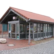 TourismusService Bruchhausen-Vilsen Kleines Tourismusbüro mit rotem Ziegeldach und Glasfenstern, umgeben von gepflasterten Flächen.