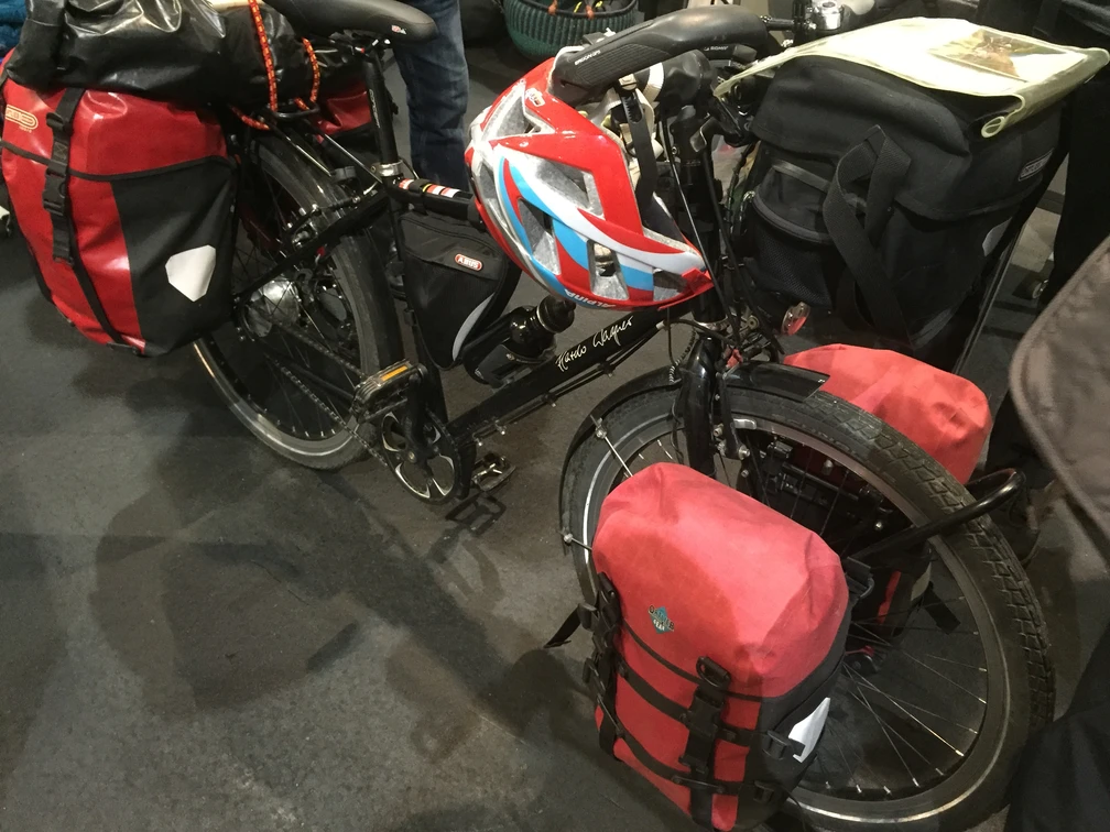 Fahrrad mit Fahrradtasche Ein schwarzes Touringrad mit roter Fahrradtasche und Helm steht gepackt auf einem Boden aus grauem Asphalt.