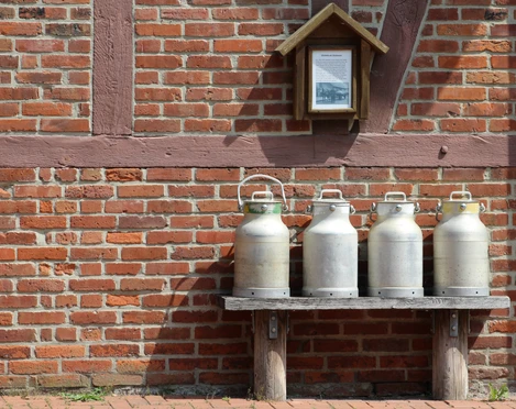 Milchkannen Vier traditionelle Milchkannen stehen auf einer Holzbank vor einer Backsteinmauer mit Fachwerk.
