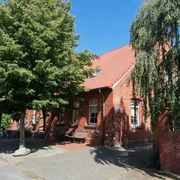 Rotes Backsteingebäude mit Tradition, umgeben von Bäumen, blauer Himmel im Hintergrund, Fahrräder seitlich.