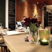 moxxa cafe die rösterei Ein eleganter Café-Innenraum mit Holztheken, auf denen rote Rosen in Glasvasen stehen, während Kerzenlicht eine gemütliche Atmosphäre erzeugt. Im Hintergrund eine Kaffeetheke.An elegant café interior with wooden counters adorned with red roses in glass vases, while candlelight creates a cosy atmosphere. A coffee bar can be seen in the background.