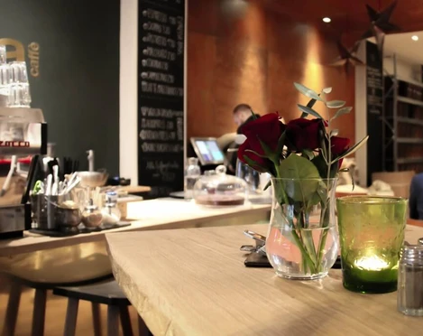 moxxa cafe die rösterei Ein eleganter Café-Innenraum mit Holztheken, auf denen rote Rosen in Glasvasen stehen, während Kerzenlicht eine gemütliche Atmosphäre erzeugt. Im Hintergrund eine Kaffeetheke.An elegant café interior with wooden counters adorned with red roses in glass vases, while candlelight creates a cosy atmosphere. A coffee bar can be seen in the background.