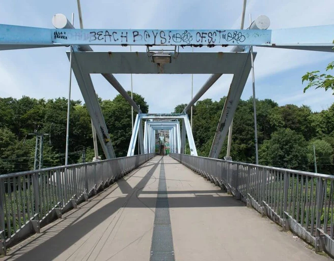 Locust bridge Fußgängerbrücke mit stählernen Trägern und künstlerischem Graffiti, umgeben von grüner Landschaft.Pedestrian bridge with steel girders and artistic graffiti, surrounded by a green landscape.
