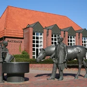 Rotes Backsteingebäude mit rotem Dach, davor Brunnen mit Figuren eines Pferdes und Menschen.
