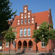 Rotes Backsteingebäude mit markantem Stufengiebel, umgeben von Bäumen und blauem Himmel.