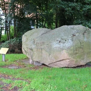 Findling Uchte Ein großer Findling aus grauem Stein ruht auf grünem Gras, umgeben von Bäumen und Büschen.