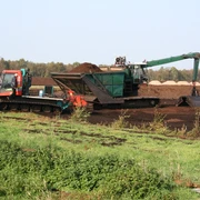 Moor Ein Torflader und ein Bagger arbeiten in einem weiten Moorgebiet unter klarem Himmel, umgeben von Vegetation.