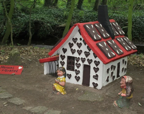 Märchenwald Pfarrbusch Das Bild zeigt ein kleines Lebkuchenhaus im Wald, verziert mit Herzformen, umgeben von Figuren.