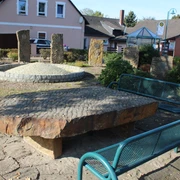 Brunnenplatz mit großen, flachen Natursteinen und modernen Bänken, umgeben von Sträuchern und Bäumen.