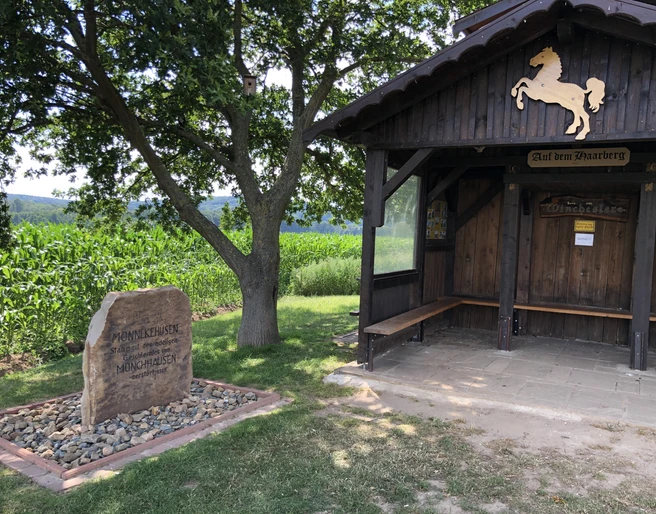 Aussichtspunkt Haarberg Holzhütte mit Wappenpferd, daneben ein Gedenkstein, umgeben von einem Kornfeld und grüner Natur.