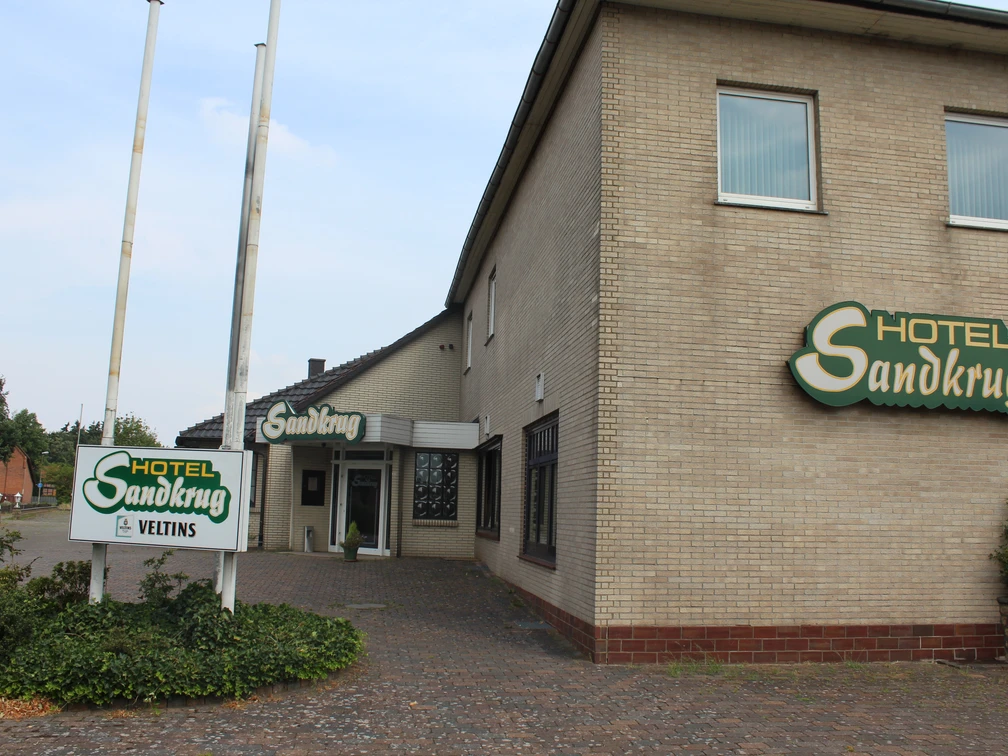 Ein großes Backsteingebäude des Hotel Sandkrug mit grünen Schildern und einem Kopfsteinpflasterweg.