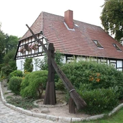 Ein historischer Ziehbrunnen steht vor einem Fachwerkhaus umgeben von Büschen und bunten Blumen.