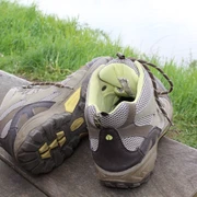 Zwei getragene Wanderschuhe stehen auf einer Holzbank vor einer grünen Wiese und einem Gewässer.