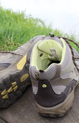 Wanderschuhe Zwei getragene Wanderschuhe stehen auf einer Holzbank vor einer grünen Wiese und einem Gewässer.