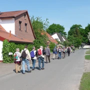 Eine Gruppe von Wanderern mit Rucksäcken spaziert auf einer Dorfstraße entlang traditioneller Häuser.
