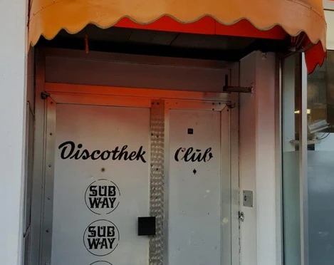 Subway Silberne Edelstahltür unter einer gelben Markise, beschriftet mit "Discothek Club" und Subway-Logo.Silver stainless steel door under a yellow awning, labeled "Discothek Club" and Subway logo.