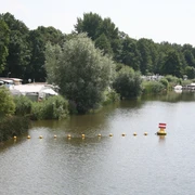 Campingplatz und Yachtclub Lahde Ruhiger See mit Segelbooten und Wohnmobilen am Ufer, umgeben von üppigen grünen Bäumen und Schilf.