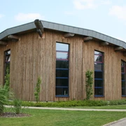 Ein modernes rundes Gebäude mit Holzfassade und markant geneigtem Metalldach, von Natur umgeben.