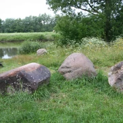 Drei große, bemooste Steine liegen in einer grünen Wiese vor einem Fluss, umgeben von Bäumen.