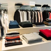 Style the brandstore Eine stilvoll eingerichtete Boutique mit gestapelten Pullovern in verschiedenen Farben auf einem Tisch und ordentlich aufgehängten Kleidungsstücken im Hintergrund.A stylishly furnished boutique with piles of sweaters in different colors on a table and neatly hung clothes in the background.