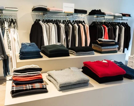 Style the brandstore Eine stilvoll eingerichtete Boutique mit gestapelten Pullovern in verschiedenen Farben auf einem Tisch und ordentlich aufgehängten Kleidungsstücken im Hintergrund.A stylishly furnished boutique with piles of sweaters in different colors on a table and neatly hung clothes in the background.