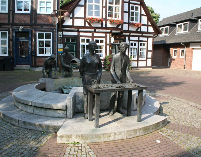 Spargelbrunnen Skulptur eines Spargelbauern mit Tafel und zwei weiteren Figuren vor historischen Fachwerkhäusern.