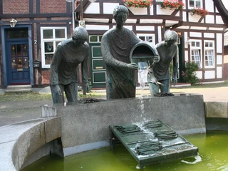Spargelbrunnen Drei Bronzefiguren schütten Spargel auf einen Gitterrost in einem halbrunden Brunnenensemble aus.
