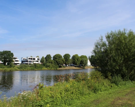 Weser Flusslauf der Weser mit grünen Ufern, modernen Gebäuden und einem Kirchturm im Hintergrund.