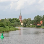 Die Weser schlängelt sich durch eine grüne Landschaft bei Nienburg; ein Kirchturm ragt im Hintergrund empor.