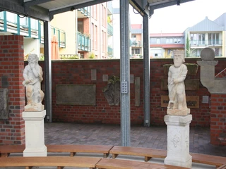 Lapidarium Nienburg Steinskulpturen auf Sockeln unter einer überdachten Fläche, umgeben von Backsteinwänden und städtischer Kulisse.
