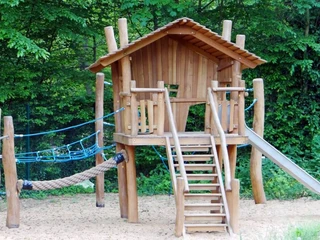 Forest swimming pool and camping Holzspielhaus mit Rutsche, Seilbrücke und Kletternetz auf Sand umrahmt von üppig grünem Wald.Wooden playhouse with slide, rope bridge and climbing net on sand surrounded by lush green forest.