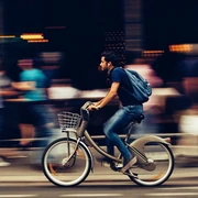 pexels-snapwire-310983(1).jpg Ein Mann fährt in rasanter Geschwindigkeit auf einem silbernen Leihfahrrad, umgeben von verschwommenen, passierenden Fußgängern einer städtischen Straße.