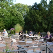 Dank Augusta Außenterrasse mit grüner natürlicher Umgebung. Menschen sitzen an Tische und unterhalten sich.Outdoor terrace with green natural surroundings. People are sitting at tables and talking.